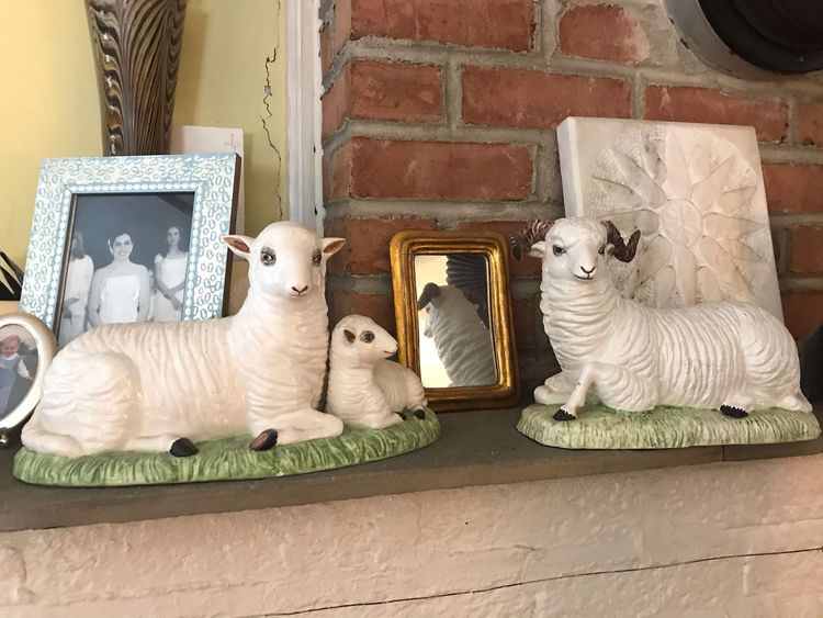 Two ceramic sheep figurines: a ram and a ewe with a lamb. The sheep are on a mantelpiece surrounded by family photos.